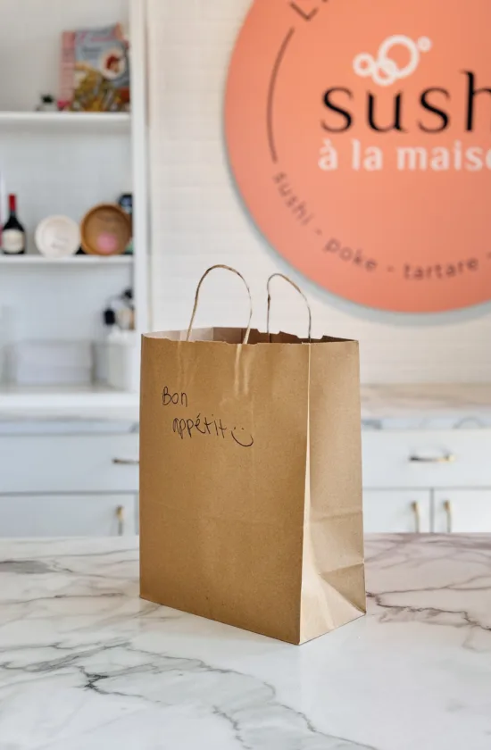 Sac en papier brun avec l’inscription “Bon appétit” posé sur un comptoir de marbre devant le logo Sushi à la maison