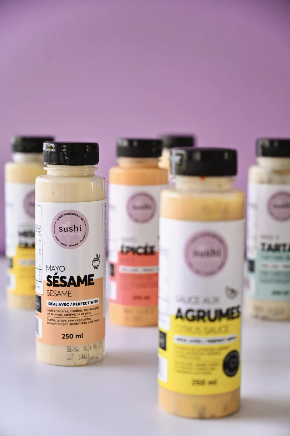 Bouteilles de sauces Sushi à la maison, dont la mayo sésame, mayo épicée et sauce aux agrumes, sur fond lilas.